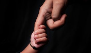 An adult holds a child's hand against a dark background